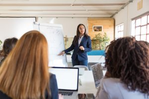 Businesswoman pointing at whiteboard during presentation. Multiethnic female colleagues looking at young speaker standing near whiteboard in meeting room. Business meeting concept